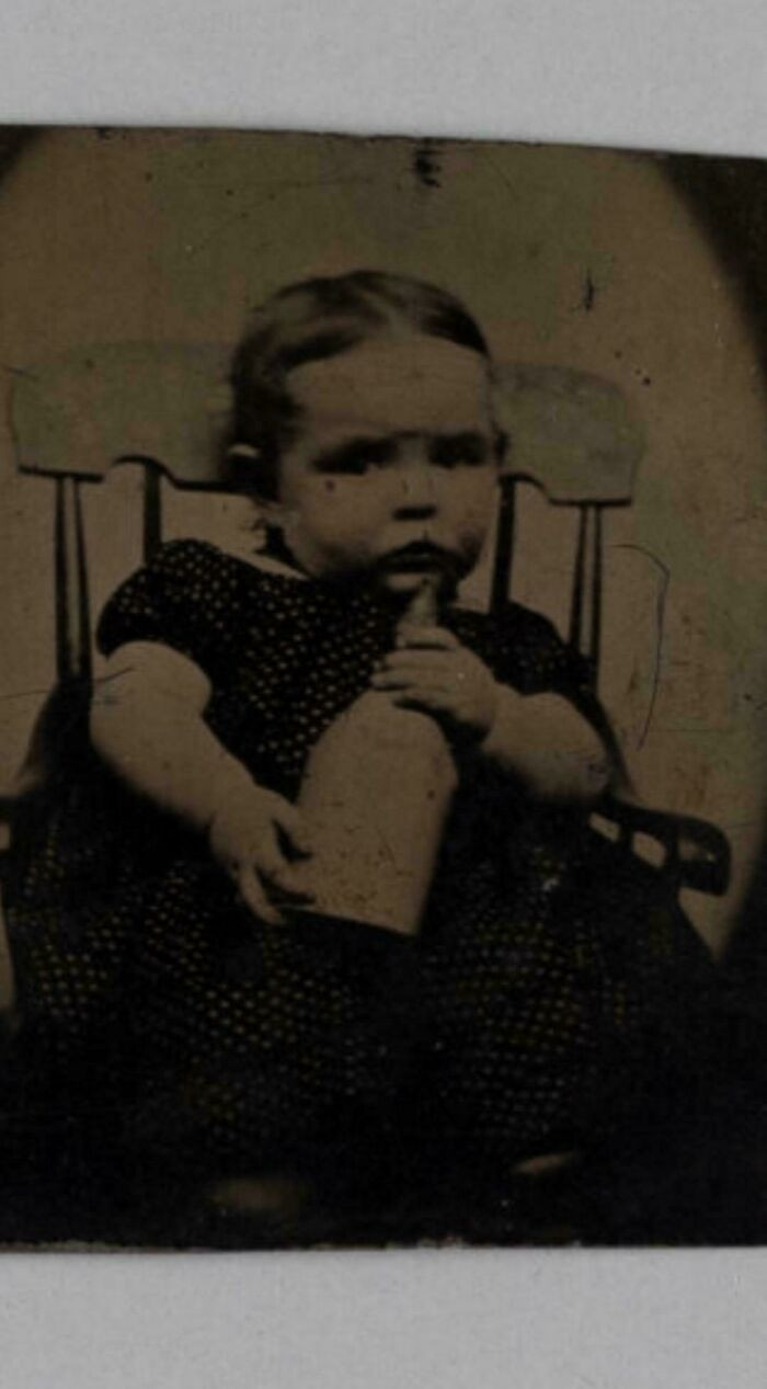 "Tintype Portrait Of A Boy Named George White Holding A Pewter Nursing Bottle, He Wears A White Dotted Dress And Sits In A Miniature "Boston Rocker", 1840-1845"