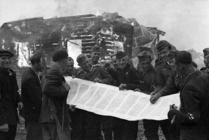 "German Soldiers and Lithuanian Auxiliary Police Laugh with a Looted Torah Scroll, Against the Backdrop of a Burning Synagogue in Occupied Lithuania, 1941."
