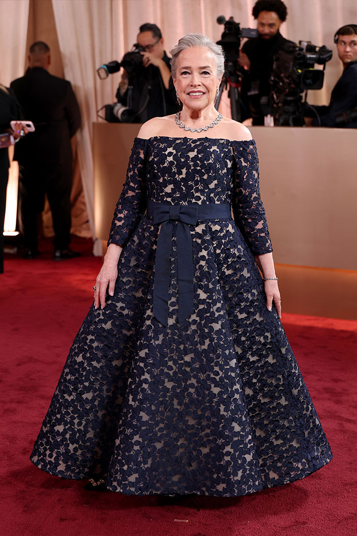 Kathy Bates wore an off-the-shoulder navy gown with a delicate foliage pattern and a bow at the waist, finishing the look with diamond jewelry and a sleek silver updo.