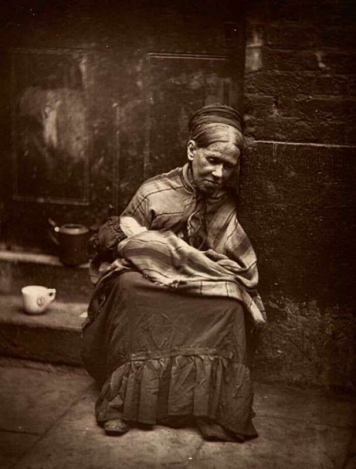 "1877. 'The Crawlers' Were The Lowest Of The British Poor. This Elderly Widow Is Sitting Outside A Tailors Shop, Holding A Baby While Its Mother Works. She Was Given A Cup Of Tea And A Slice Of Bread Daily In Return"
