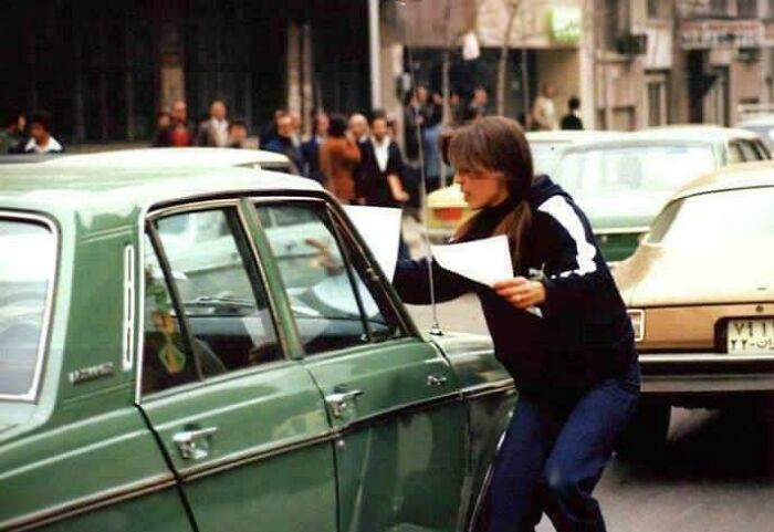 A young Iranian woman distributes anti-Shah leaflets in Tehran during the Iranian Revolution, capturing a moment of quiet resistance in 1979.