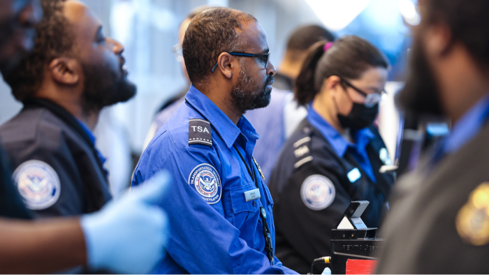 Airport security area showing travelers and TSA staff amid DHS funding lapse delays.