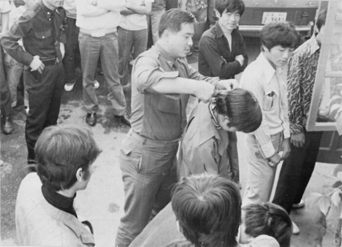 "A South Korean Soldier Forcibly Cuts A Young Man's Hair In Front Of Others During A Nationwide Crackdown On Men With Long Hair And Women Wearing Short Skirts In South Korea - 1970s"