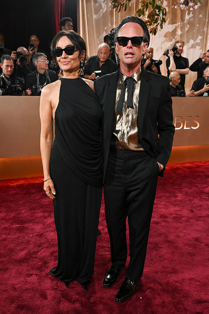 Nadia Conners wore a sleek, sleeveless, black wrap dress with a high neckline and gentle side draping. Goggins complemented her look with a black suit, a shimmering gold shirt, and a slim black tie.