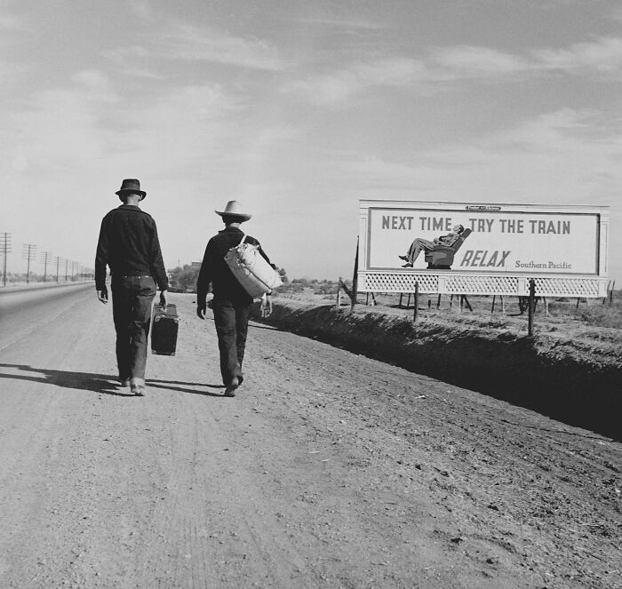 Two pedestrians walk down a roadside path in 1937, passing a billboard that reads, “Next time, try the train. Relax.”