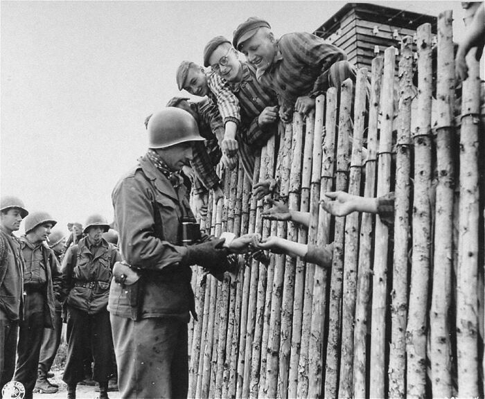 U.S. Army Corporal Larry Matinsk Puts Cigarettes Into The Extended Hands Of Newly Liberated Prisoners Behind A Stockade In The Allach Concentration Camp, April 30, 1945
